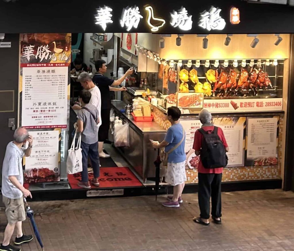 Hong Kong locals gather outside a restaurant that has roast goose and chicken hanging from hooks in a window.