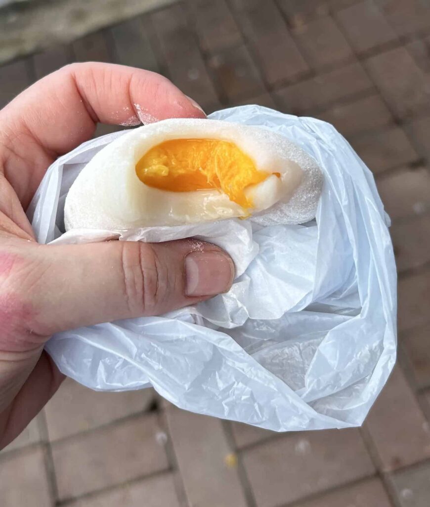 A hand holding a mango mochi with a bite taken out of it, revealing a golden mango center.