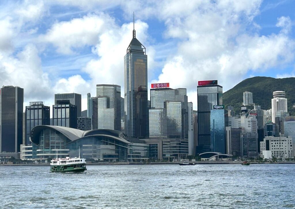 The Star Ferry crosses a calm Victoria Harbour. The Hong Kong Convention centre and other skyscrapers are in the background. The sky is blue with white clouds.
