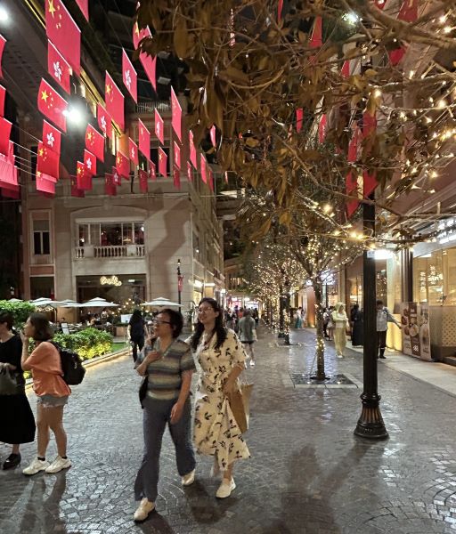 Two smiling women walk along the cobblestone Lee Tung avenue at Night. Chinese flags are string up and lights are in the trees.