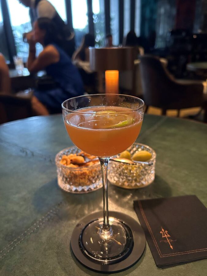 An orange drink in a cocktail glass. Two glass containers of olives and nights, In the foreground a waiter takes a the drinks order of a party of three people.