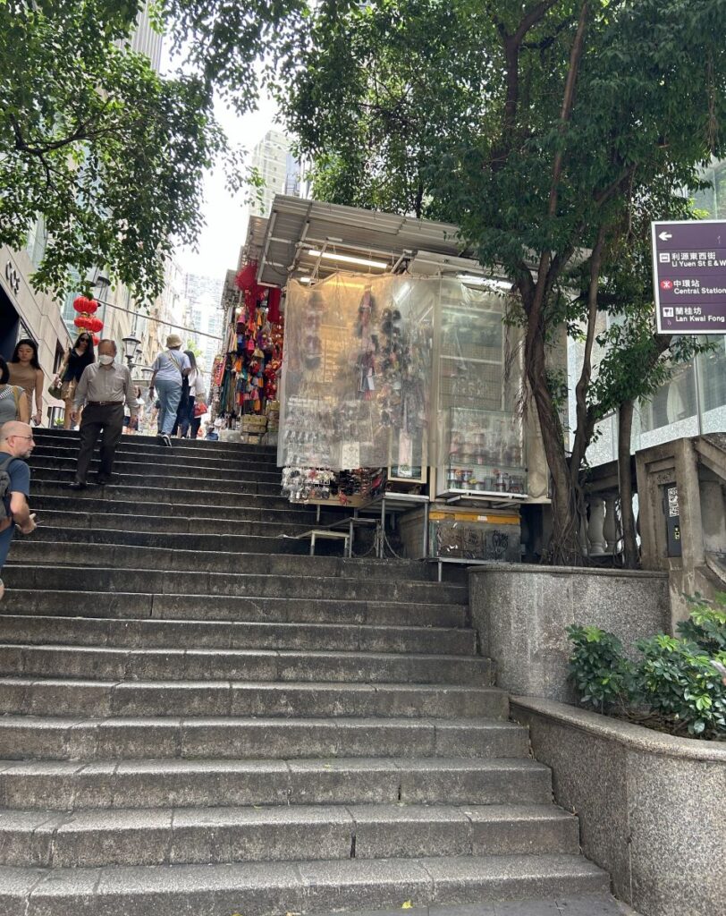 Stone steps leading up to market stalls. A canopy of trees provides some shade.