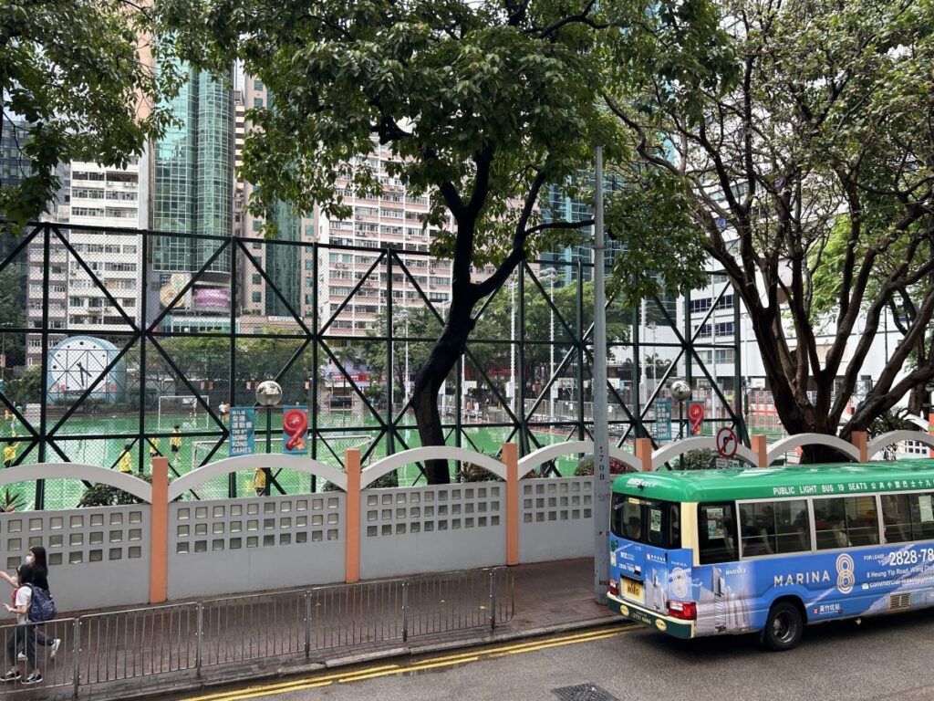 A mini bus is parked outside the Outdoor Wan Chai Basketball courts. Boys are playing basketball and the court is wet from rain fall.