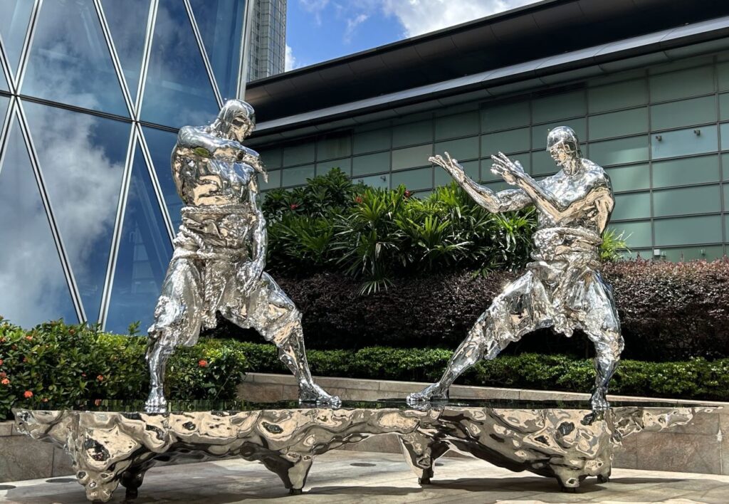 Two futuristic silver statues in martial arts stances stand on an abstract metallic base outside a modern Hong Kong building, symbolizing the city’s cultural legacy of kung fu and action cinema—key elements of Hong Kong history.