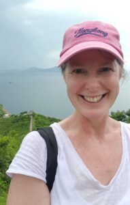 Smiling woman in a pink "Hong Kong" baseball cap and white shirt takes a selfie during a solo hike, with lush green hills and the sea stretching into the misty distance behind her. She carries a black-strapped backpack, and the sky is overcast, adding a moody vibe to the scenic backdrop.