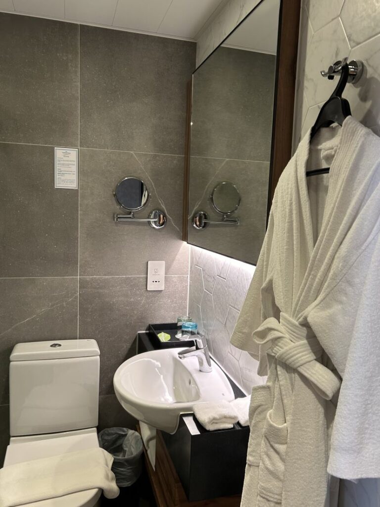 Compact hotel bathroom with modern fixtures at the Harbourview Hotel, featuring a wall-mounted sink, toilet, and a large mirror with a mounted magnifying mirror. A white bathrobe hangs on a hook beside the sink, and folded towels, drinking glasses, and amenities are neatly arranged on the counter.